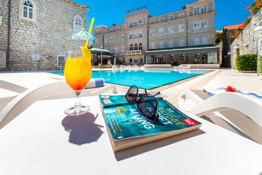 ein Getränk und ein Buch auf einem Tisch neben einem Pool in der Unterkunft Hotel Lapad in Dubrovnik