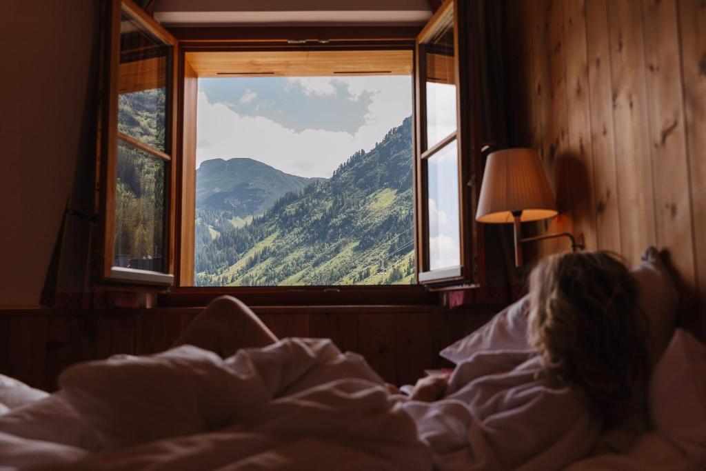 a woman laying in bed looking out a window at Hotel Aurora in Lech am Arlberg