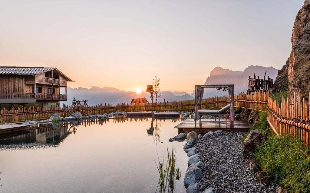 a view of a lake with a house and the sunset at Hotel Goldknopf in Alpe di Siusi