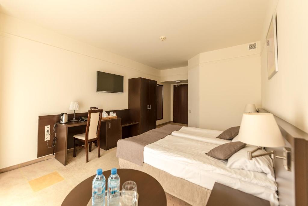 Bright hotel bedroom with two beds, bedside lamps, desk area and water bottles on table.