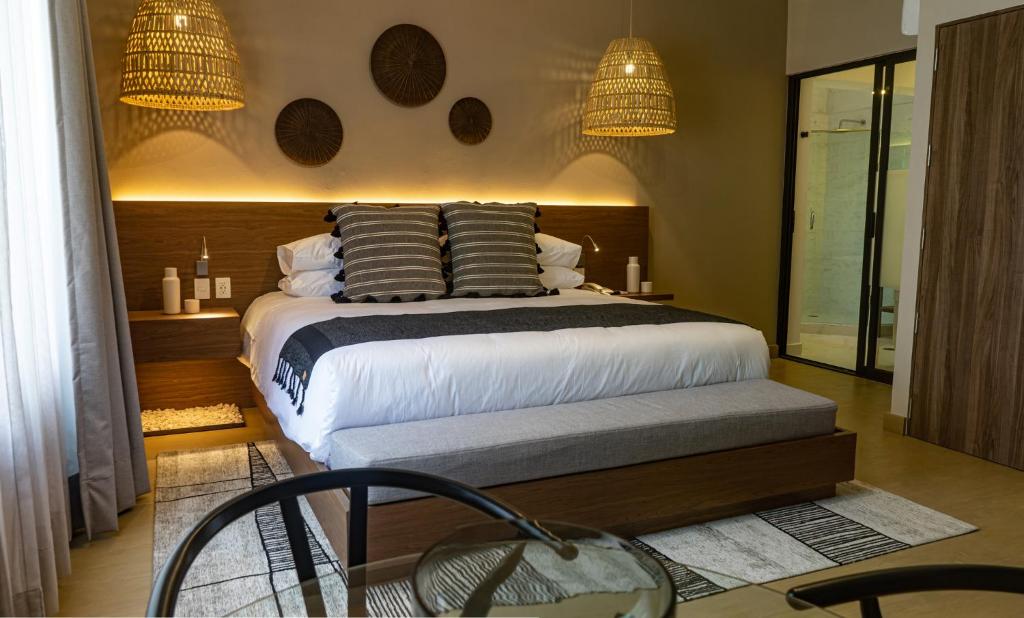 A large bed with a foot bench, woven pendant lights and a glass-fronted bathroom doorway.