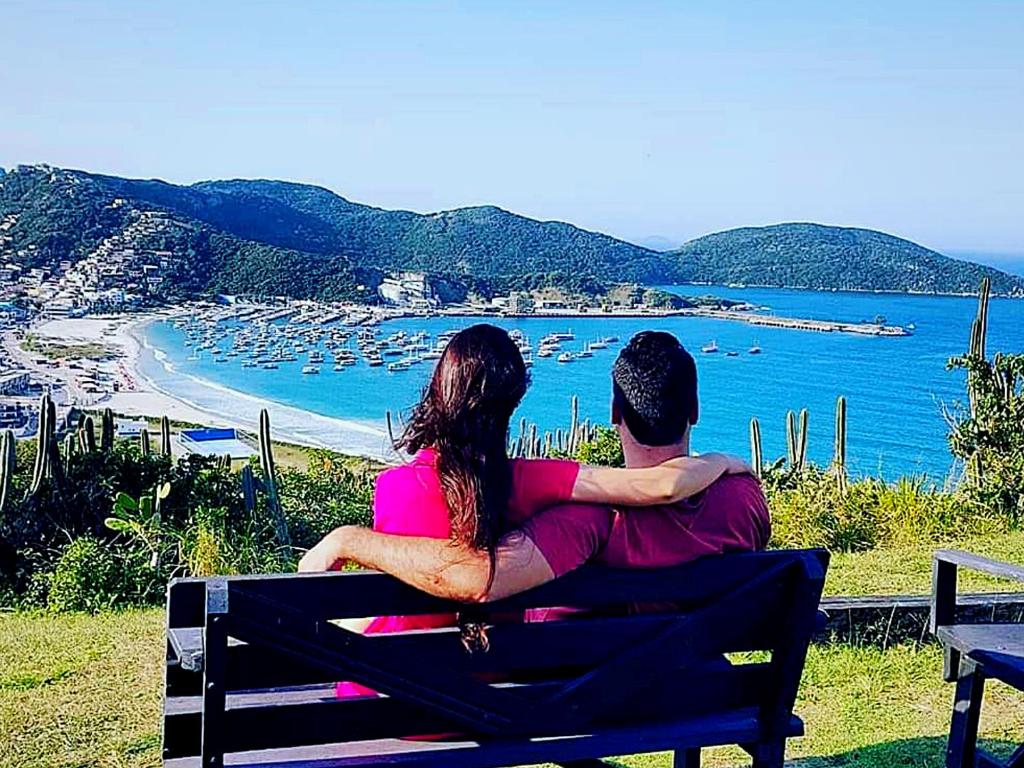 a man and woman sitting on a bench looking at a beach at Pousada Tanto Mar in Arraial do Cabo