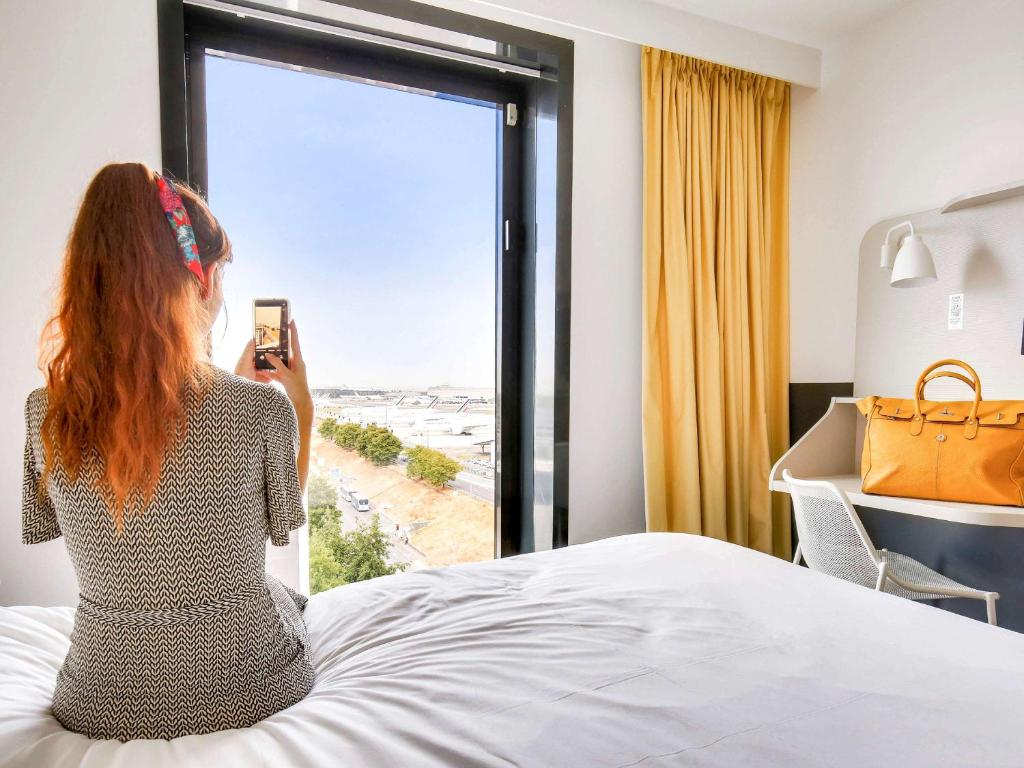 a woman standing in front of a window looking at her phone at ibis Styles Paris Charles de Gaulle Airport in Roissy-en-France