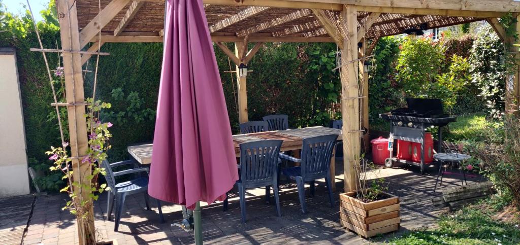 een houten pergola met een tafel en stoelen en een roze parasol bij L'Alisier Chantant in Bourron-Marlotte
