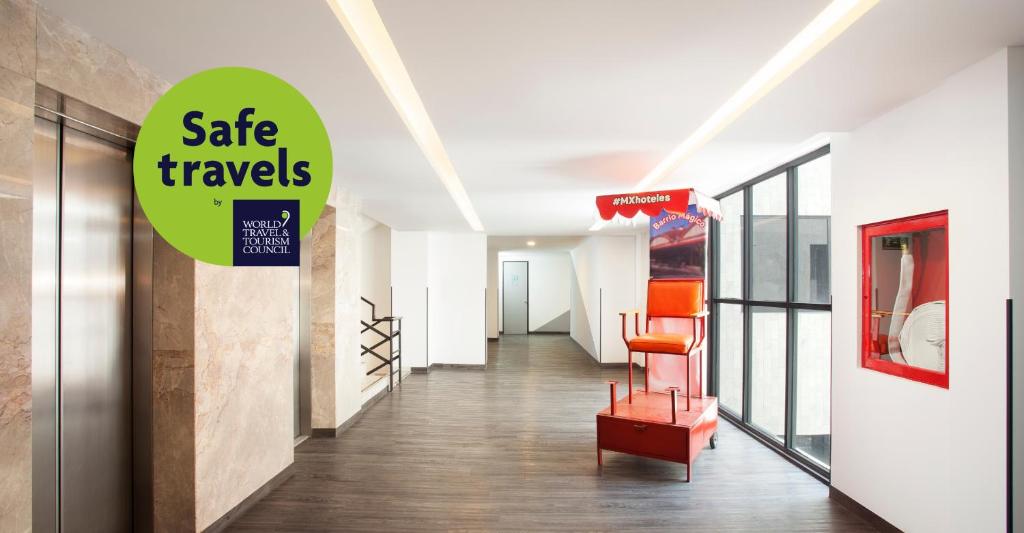 A bright hotel corridor with an elevator, glass walls, and a red sculptural chair.