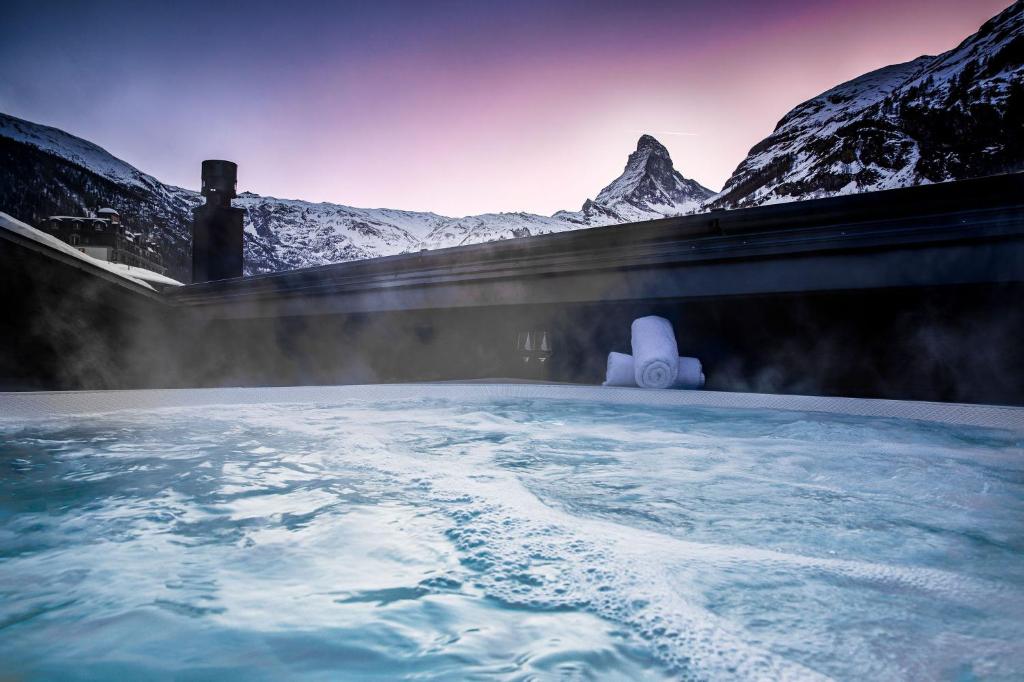 a hot tub with snow covered mountains behind it at Hotel ZERMAMA Zermatt in Zermatt