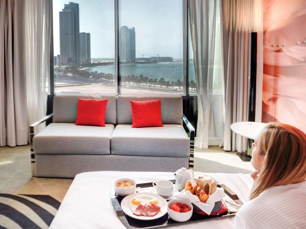 a woman sitting in a hotel room with a tray of food at Novotel Sharjah Expo Centre in Sharjah
