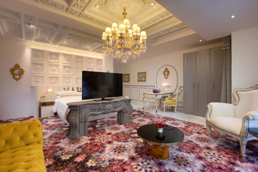 An ornate living room with a television, chandelier, sofa seating and a bed visible beyond.