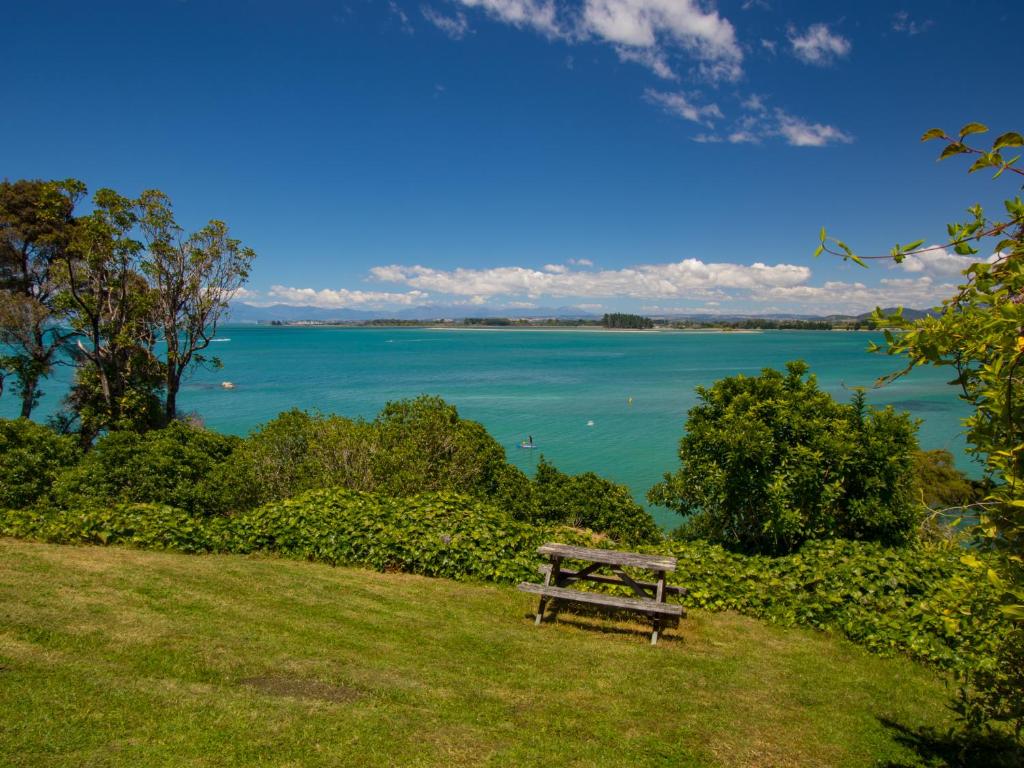 a wooden bench sitting on a grassy hill overlooking a lake at Bay Breeze - Stephens Bay, Kaiteriteri Home in Kaiteriteri