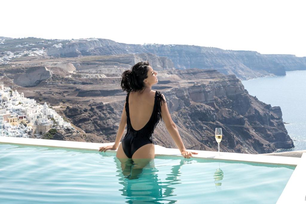 a woman sitting in a infinity pool looking out over the ocean at Mythical Blue Luxury Suites in Fira