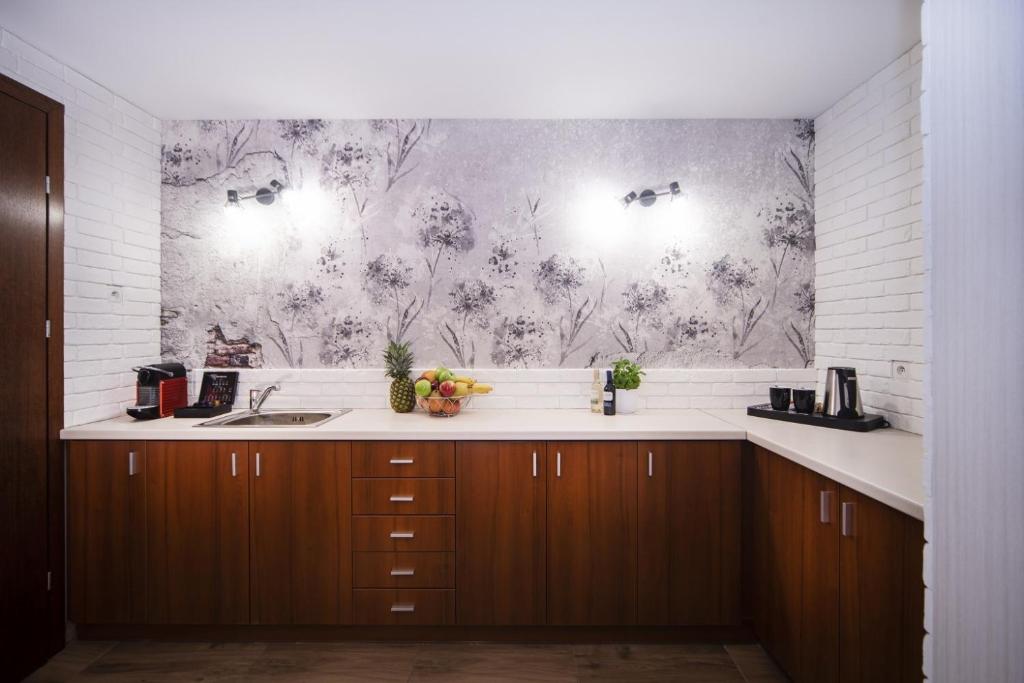 Modern kitchenette with wooden cabinets, sink, coffee machines, fruit bowl, white brick walls, floral wallpaper.