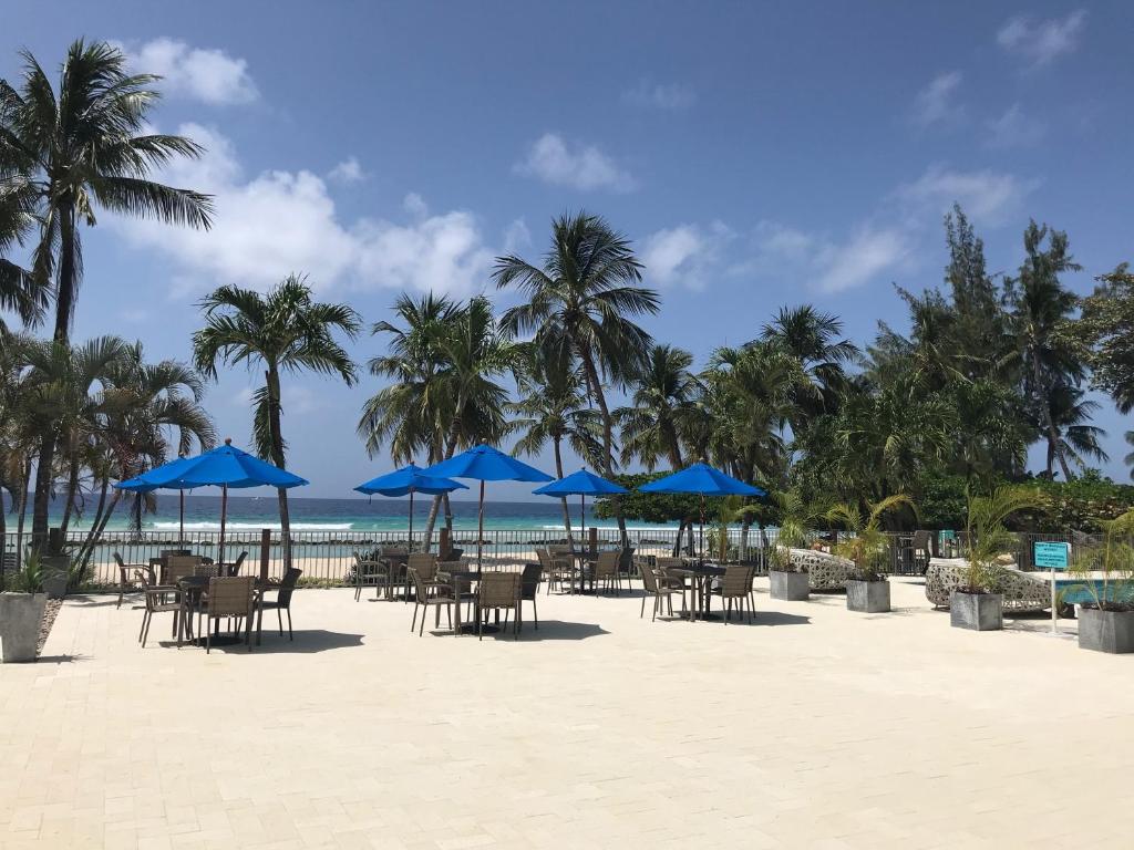 a beach with tables and chairs and blue umbrellas at Coconut Court Beach Hotel in Bridgetown