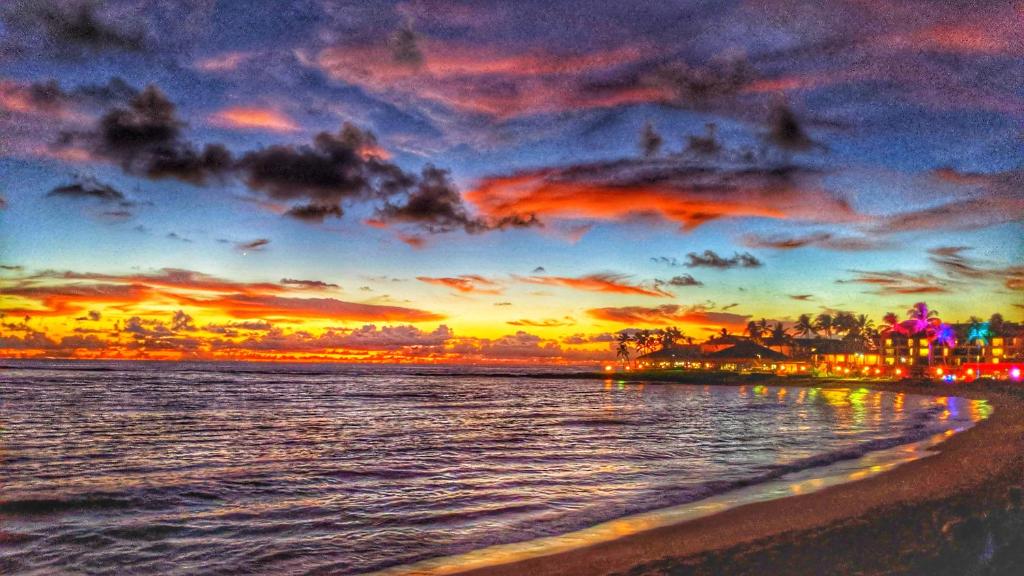 a sunset on the beach with a city in the background at ALOHA - Poipu Beach Vacation Condo in Koloa