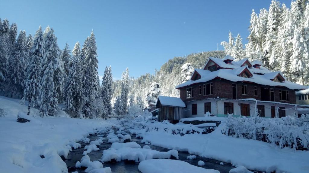 a lodge covered in snow next to a river and trees at Shiva Valley Riverside Home Stay in Jibhi