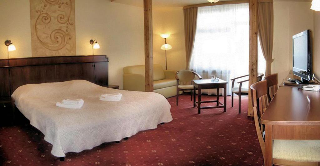 Generous hotel suite with king bed, seating area, desk, television, warm lighting and red carpet.