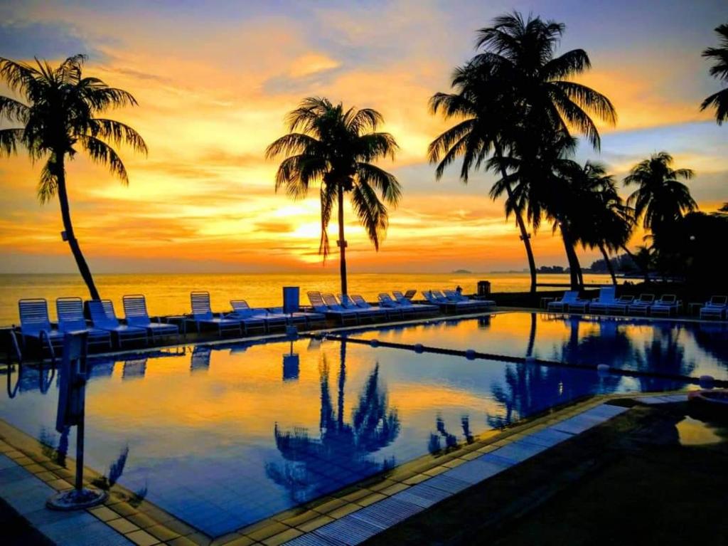 a swimming pool with palm trees and a sunset at Tanjung Tuan Beach Regency - Hermis Theme in Kampong Baharu