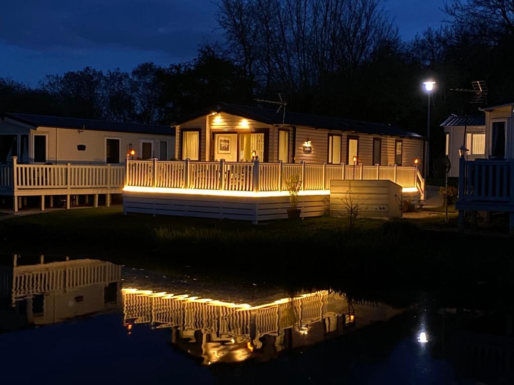 una casa con luces en el agua por la noche en Auroras Dream Lodge - Hoburne Cotswolds Holiday Park en Cirencester