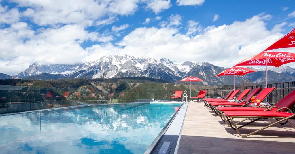 a swimming pool with chairs and umbrellas and mountains at Hotel Erlebniswelt Stocker in Schladming