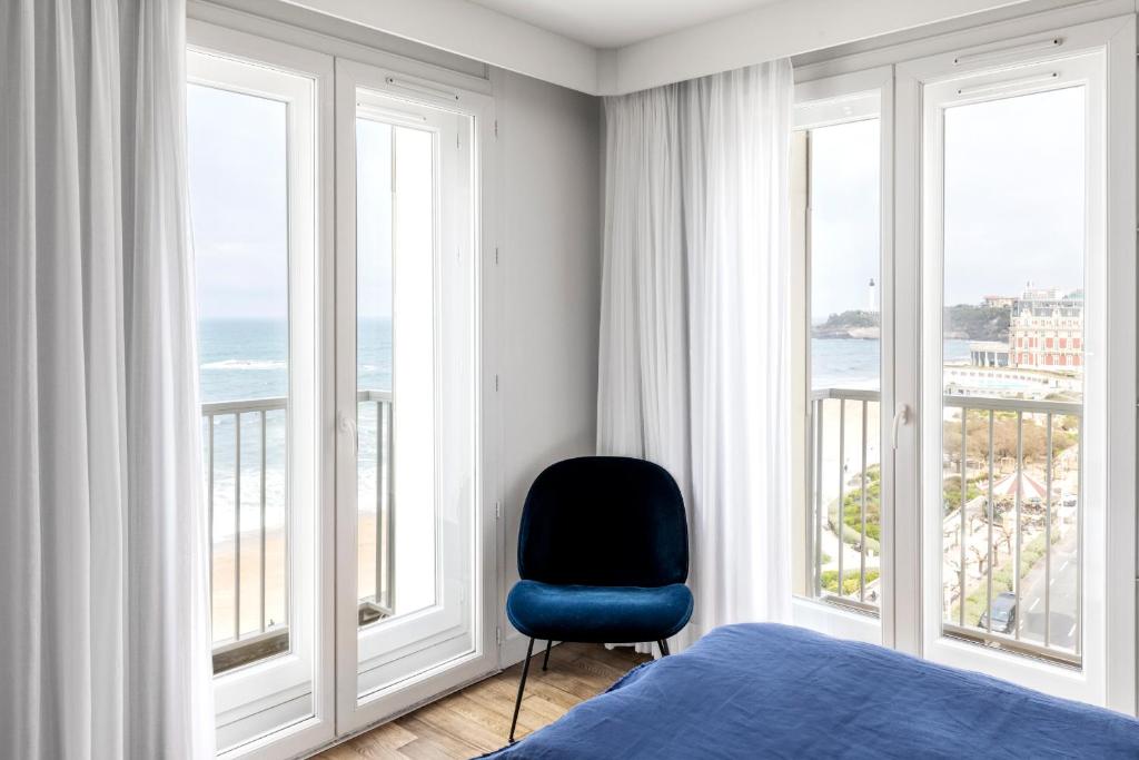 a bedroom with a blue chair in front of windows at Hôtel Le Windsor Biarritz in Biarritz