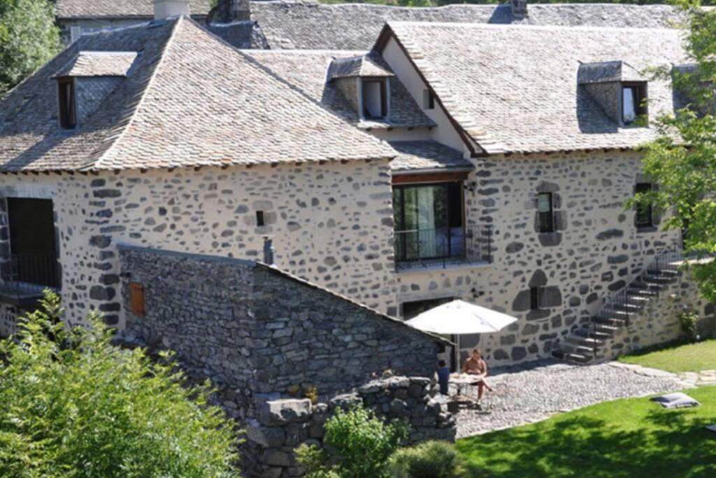 a woman sitting under an umbrella in front of a stone house at Gite aux Caprices d'Aubrac in Laguiole