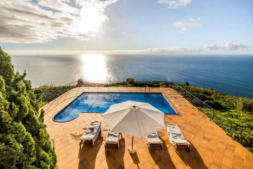 een zwembad met een parasol en stoelen en de oceaan bij Secluded Sunset Villa on Cliff & 180 Degree Ocean Views in Fajã da Ovelha