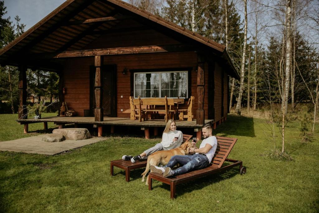 two people and a dog sitting on a bench in front of a cabin at Kalniaus-Sodyba in Barkellai
