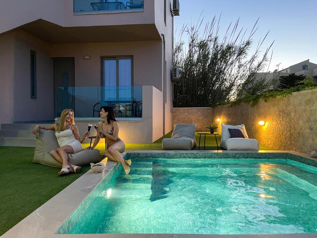 two women are sitting next to a swimming pool at Aegean Breeze Luxury Apartments next to the sea in Maleme