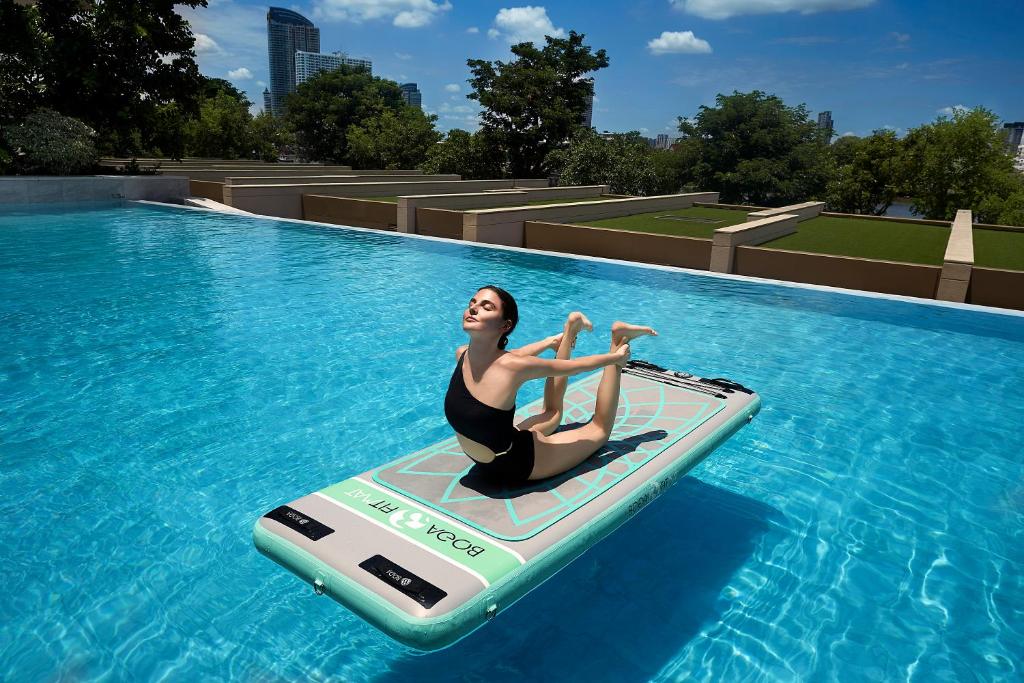 A rooftop-looking blue pool with a guest using a paddleboard and a skyline beyond.