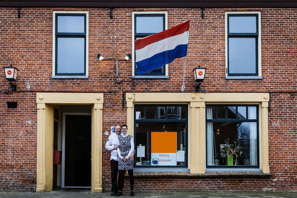 Herberg restaurant Molenrij في Kloosterburen: شخصين واقفين أمام مبنى من الطوب مع العلم