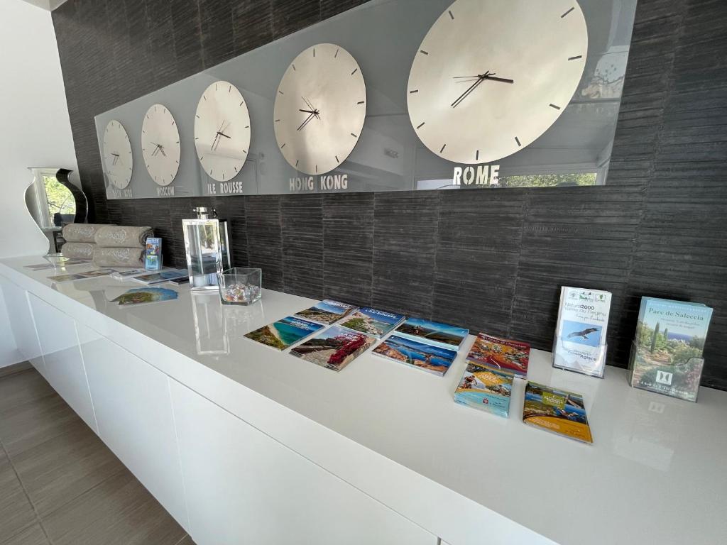a white counter with clocks on the wall and books at Hôtel L&#39;Escale Coté Port in LʼÎle-Rousse