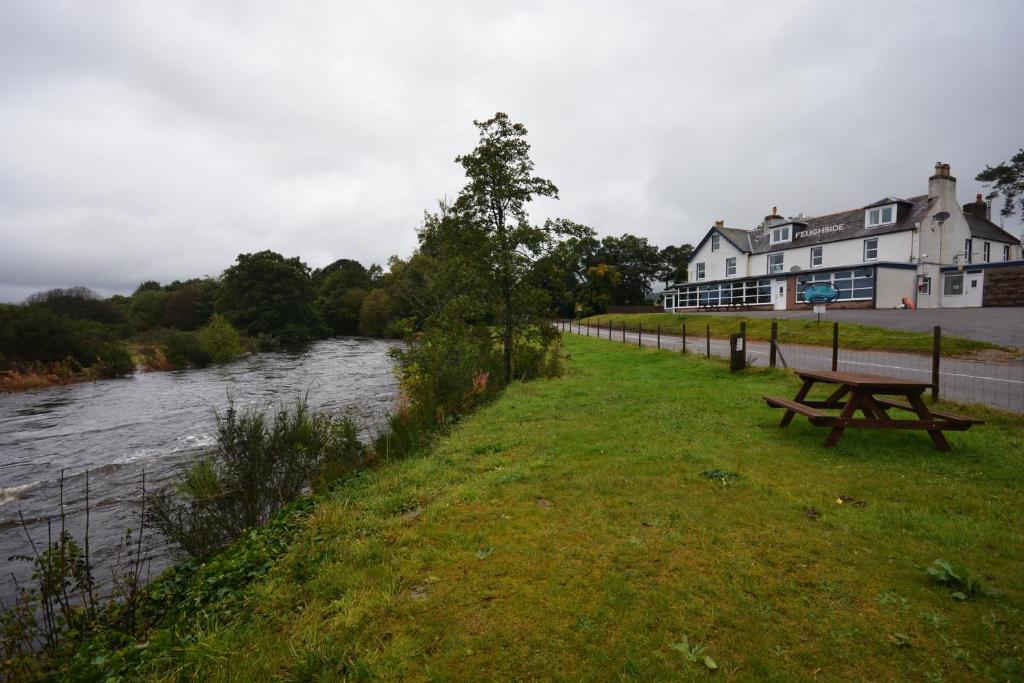 a park bench next to a river with a building at Feughside in Strachan