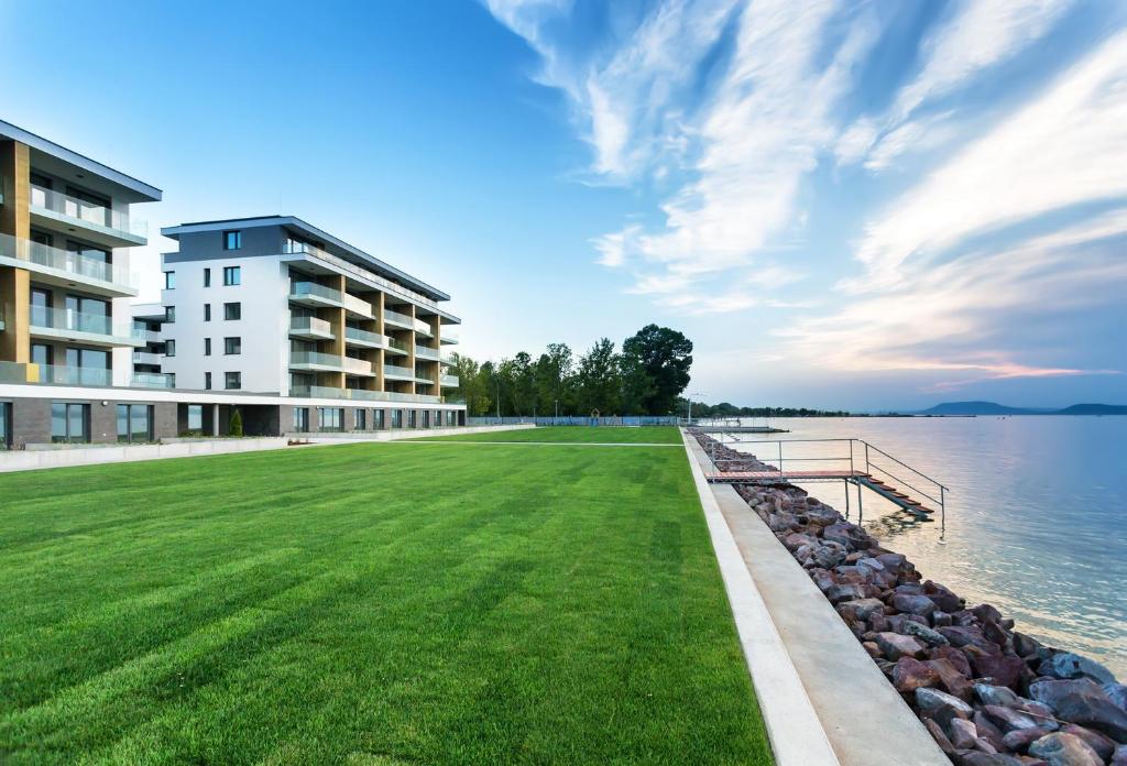 a large grassy field in front of a building next to the water at Vízpart Hullám Apartman in Balatonlelle