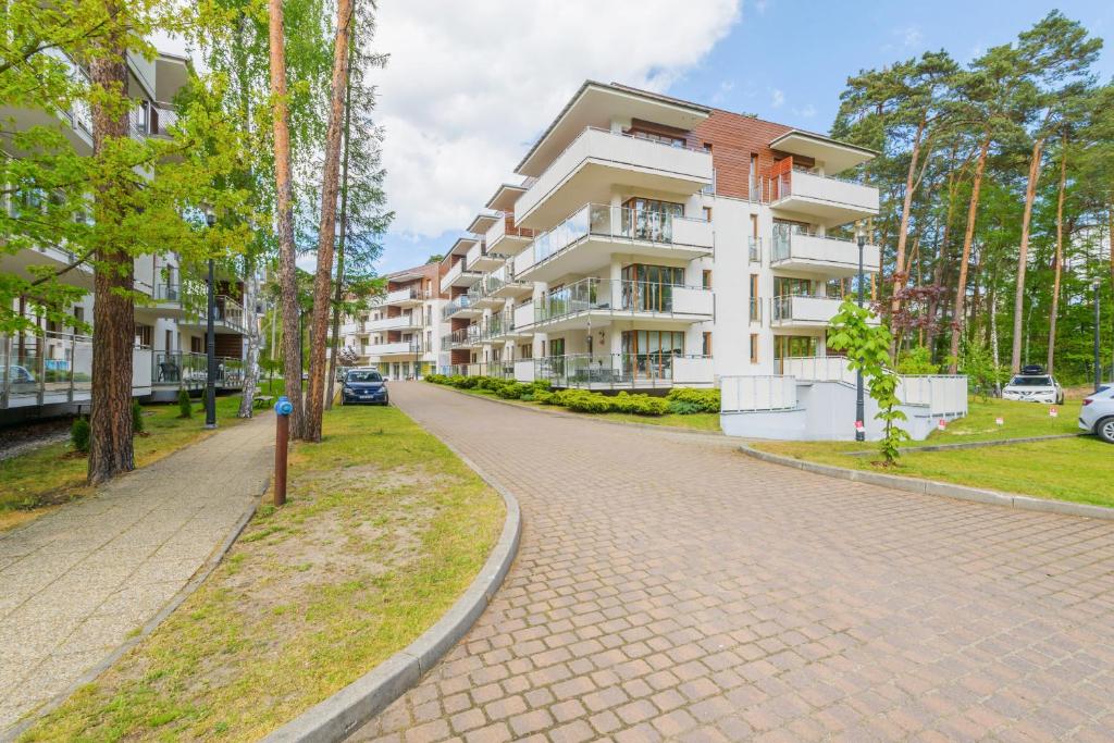 Modern white apartment buildings along curved cobblestone driveway, surrounded by pine trees and parked cars.