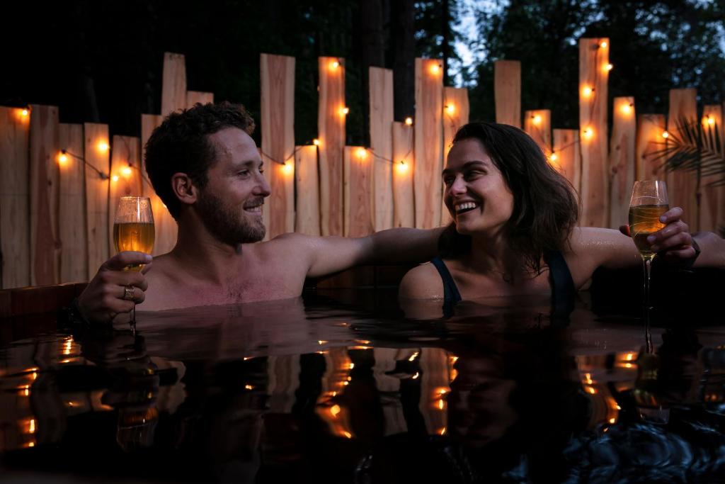 un hombre y una mujer en una piscina con copas de vino en Bain de forêt - Lodges Cosy & Spa en Neufchâtel-Hardelot