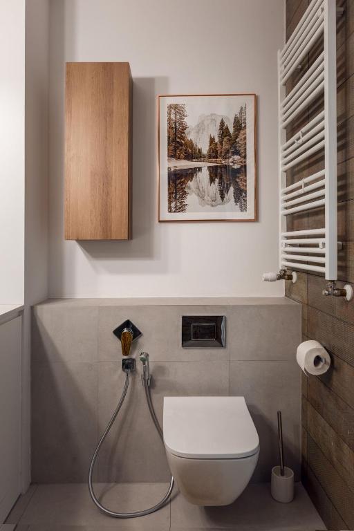 Wall-mounted toilet and bidet spray in compact modern bathroom with wooden cabinet, towel radiator, framed nature print.