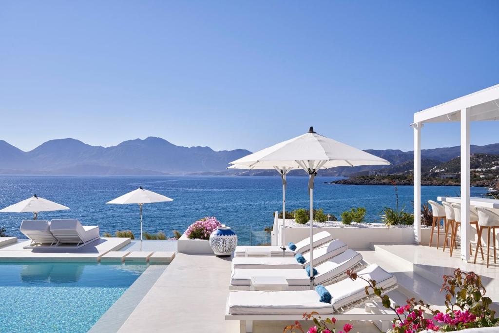 a pool with chairs and umbrellas and the ocean at The Island Concept Luxury Boutique Hotel Heated Pool in Agios Nikolaos