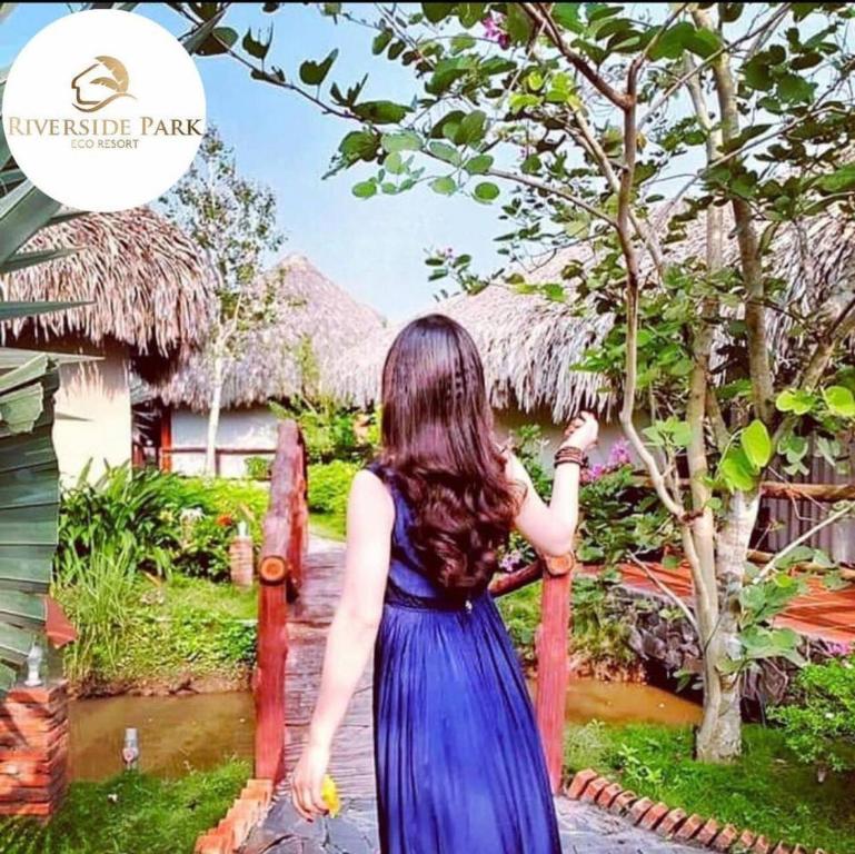 a woman in a blue dress is standing next to a tree at Riverside Park Eco Resort in Vĩnh Long