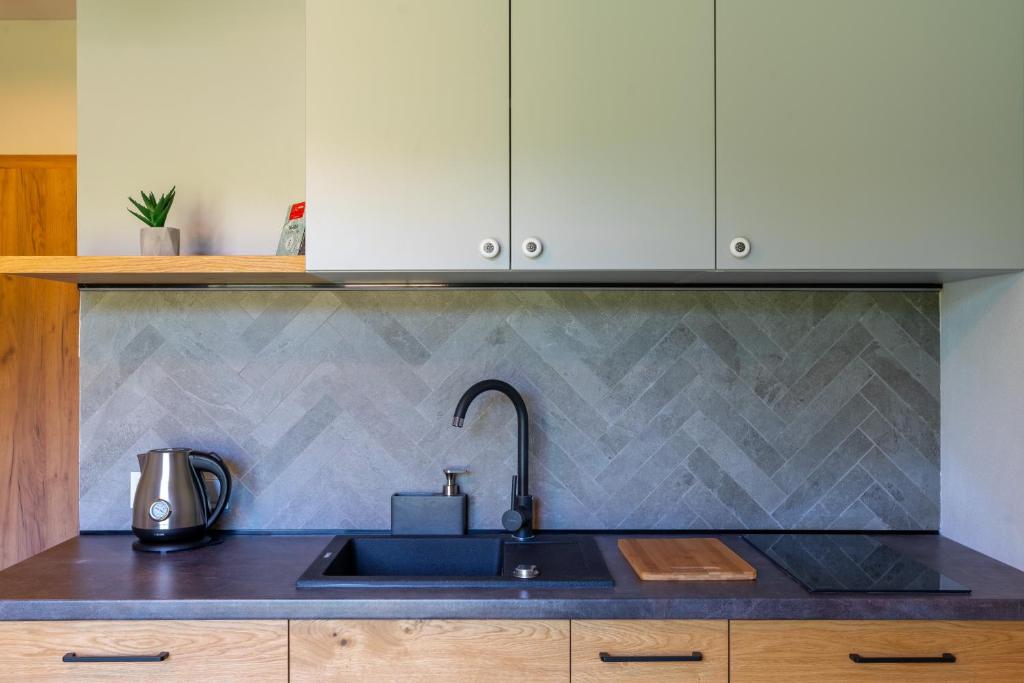 Modern kitchenette with black sink and faucet, kettle on counter, herringbone stone backsplash, warm wood cabinets.
