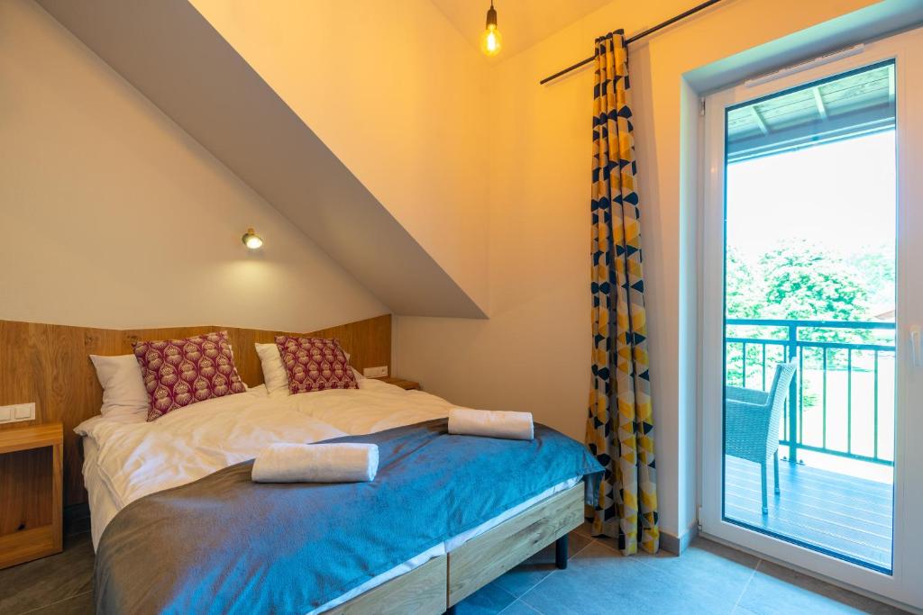 Bright attic bedroom with balcony door, patterned yellow curtains, double bed topped with rolled towels, wicker balcony chair visible.