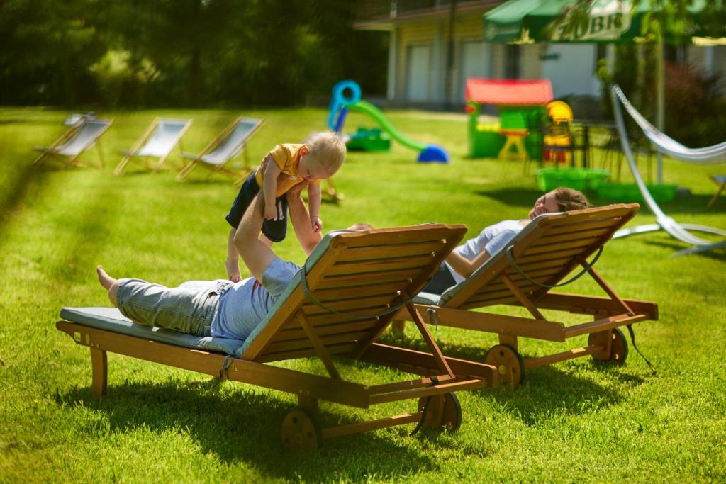 Parent lying on wooden sun lounger on sunny lawn lifting toddler playfully; partner relaxing nearby.