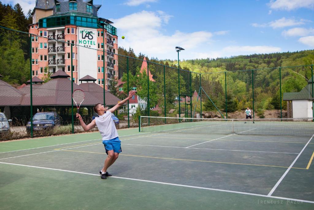 Tennis player serving on outdoor court with hotel and wooded hills in background, active scene