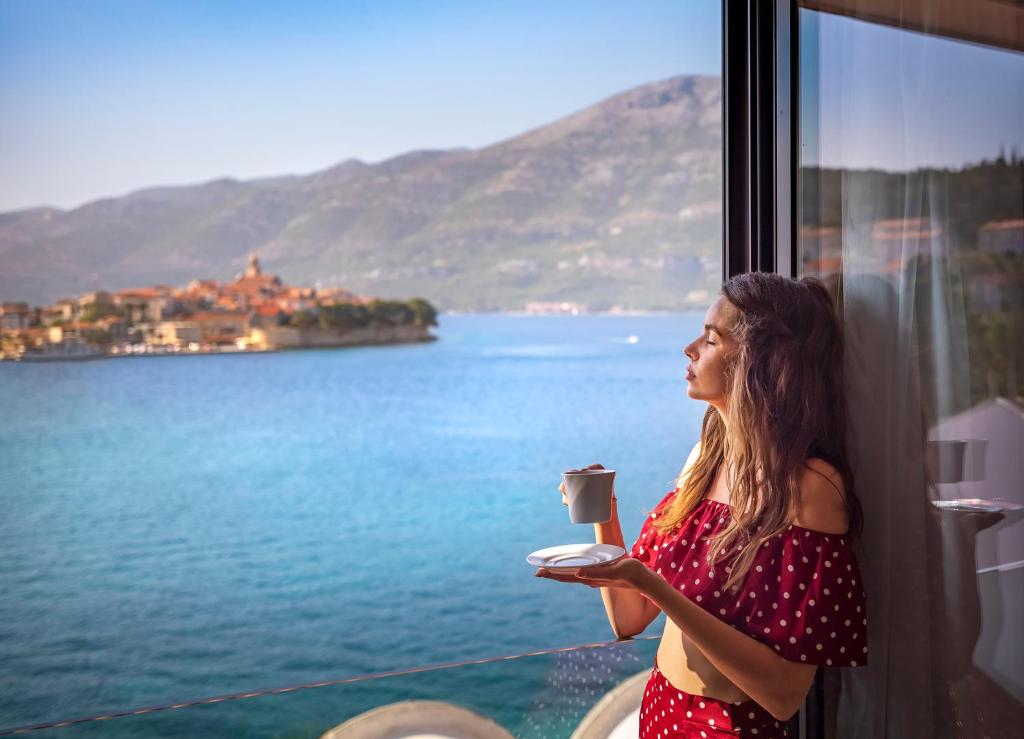 a woman holding a cup of coffee looking out a window at Aminess Liburna Hotel in Korčula