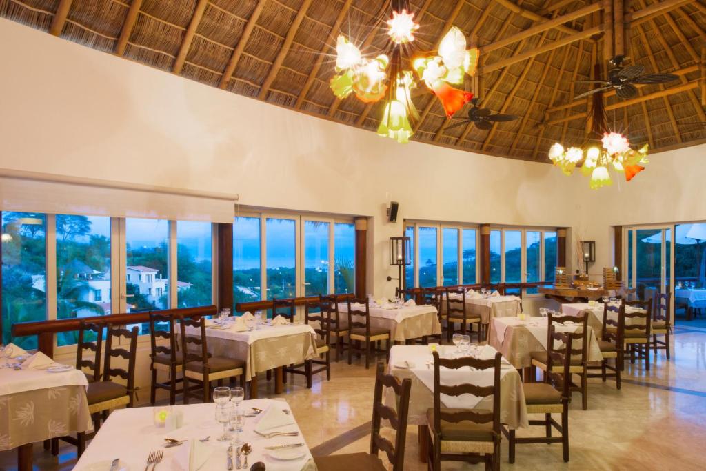 Dining room with set tables, wooden chairs, thatch ceiling, and panoramic windows overlooking landscape.