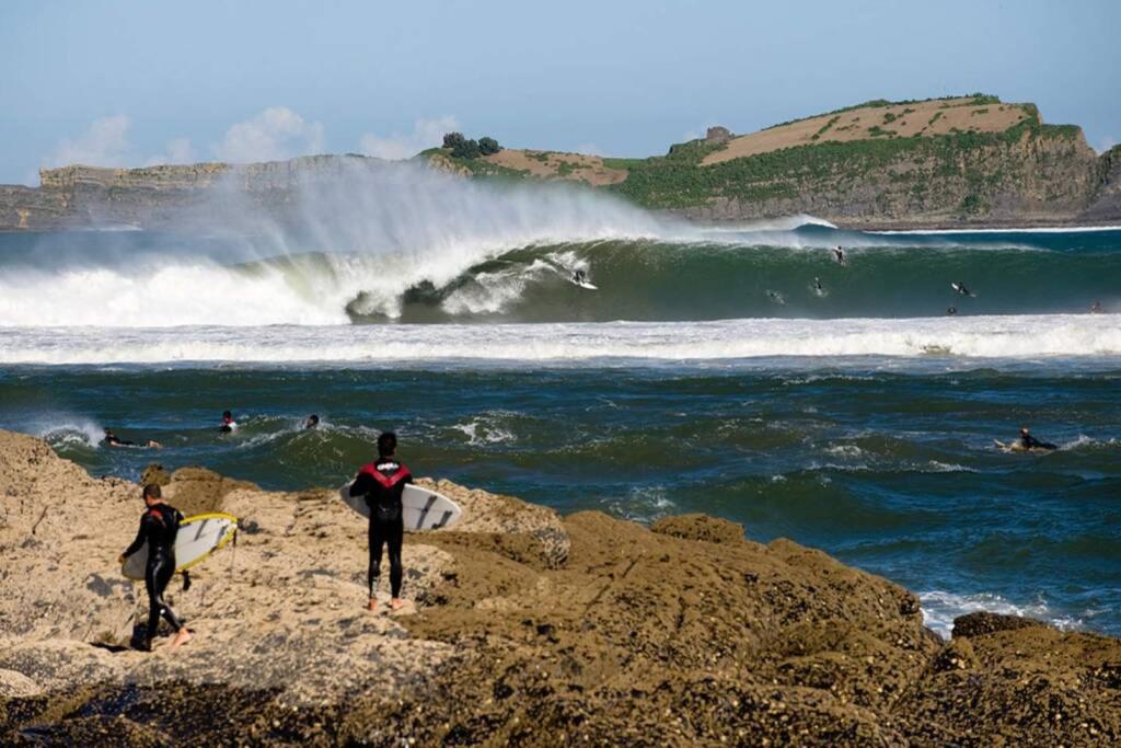 deux surfeurs sont debout sur les rochers à la vue d'une vague dans l'établissement Apartamento en el centro de Mundaka EBI646, à Mundaka