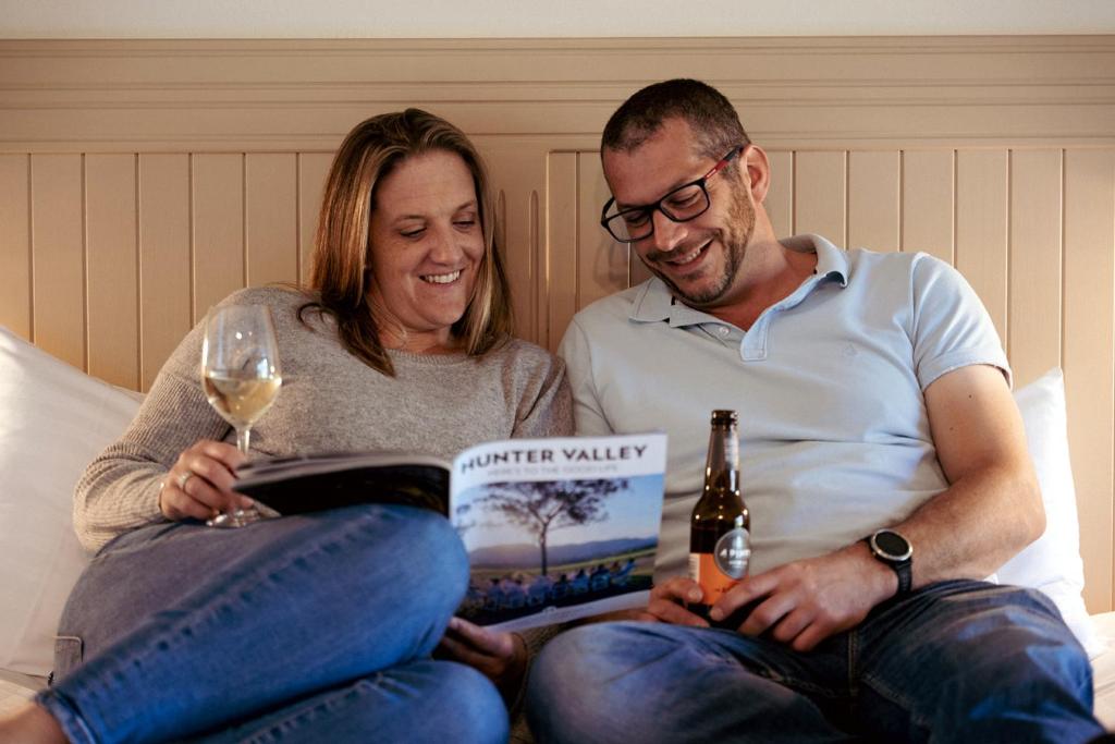um homem e uma mulher sentados numa cama com um livro e um copo de vinho em Hunter Valley Resort, Hunter Farm Adventure Centre & 4 Pines at the Farm em Pokolbin