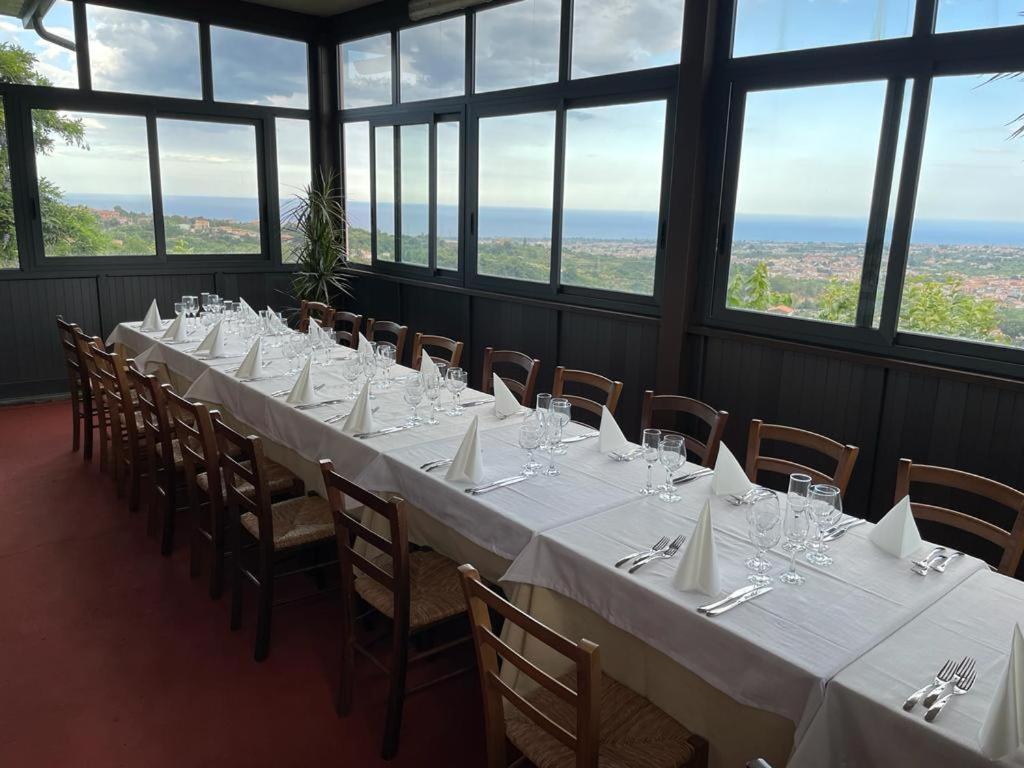 uma mesa longa com mesas e cadeiras brancas num quarto com janelas em Il noceto dell'Etna em Giarre