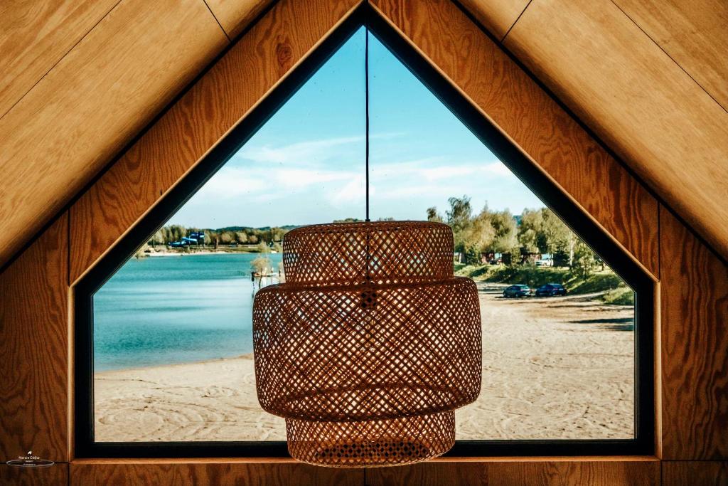 Woven pendant lamp framed by triangular window, sandy beach and lake visible outside.