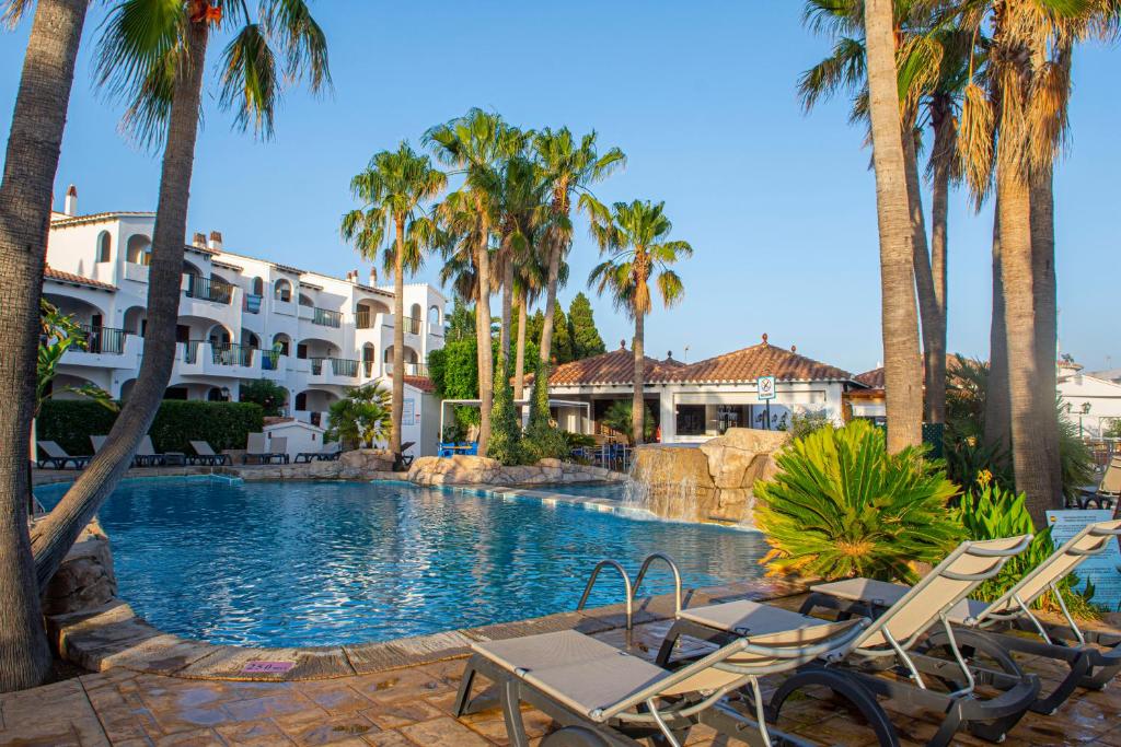 a resort swimming pool with chairs and palm trees at Siesta Mar Apartamentos in Cala'n Porter