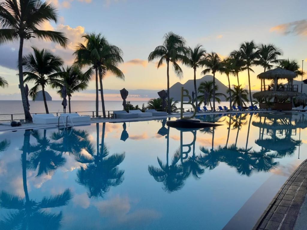 uma piscina com palmeiras e o oceano ao pôr do sol em Diamantchery - Vue mer avec Deck privatif em Le Diamant