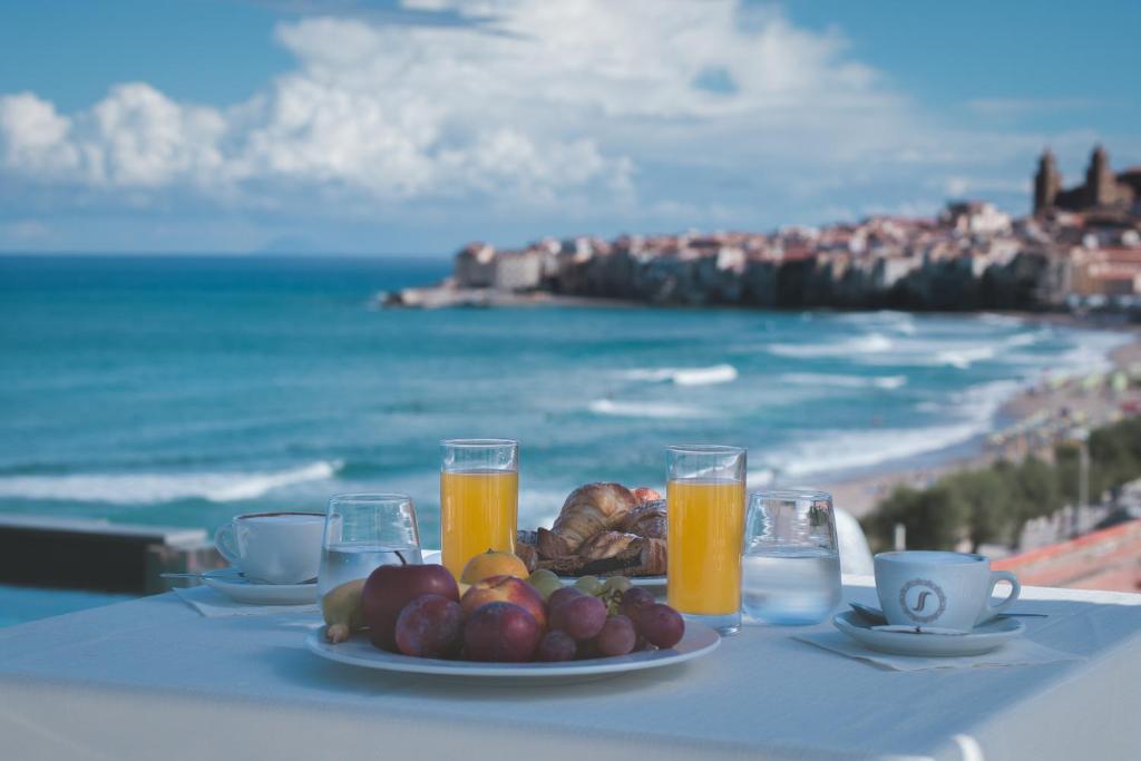 a table with a plate of fruit and juice and the ocean at Sunset Hotel in Cefalù